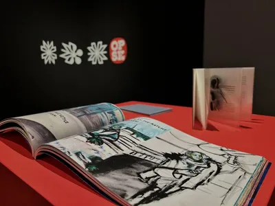 Na czerwonym stole leży kolorowa, otwarta książka o sztuce, a obok niej stoi pionowo kolejna książka. W tle widać czarną ścianę z białymi graficznymi motywami kwiatowymi oraz czerwone okrągłe logo z napisem „OPSIZ.”.